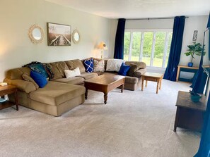 Ein Wohnzimmer mit Ecksofa, Couchtisch, Beistelltisch und einem Fenster mit Vorhängen.