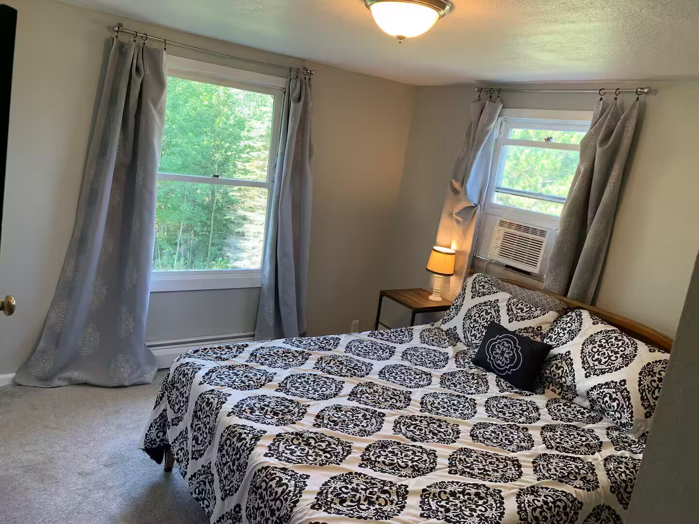 A bedroom with a bed, two windows with curtains, a bedside lamp, and an air conditioning unit.