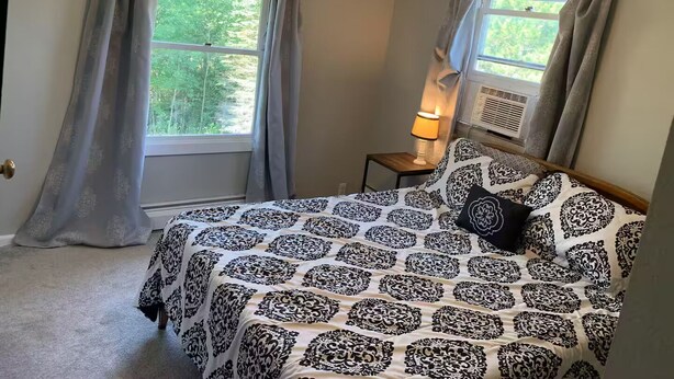 Ein Schlafzimmer mit einem Bett, zwei Fenstern mit Vorhängen, einer Nachttischlampe und einer Klimaanlage.