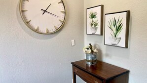 Ein Zimmer mit einer runden Wanduhr, einem kleinen Holztisch mit Blumen in einer Vase und zwei gerahmten Pflanzenbildern an der Wand.