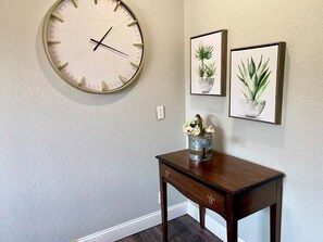 Ein Zimmer mit einer runden Wanduhr, einem kleinen Holztisch mit Blumen in einer Vase und zwei gerahmten Pflanzenbildern an der Wand.