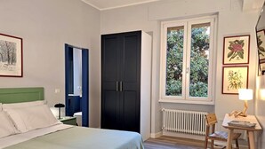 Quarto com uma cama, mesa de cabeceira, escrivaninha com cadeira, janela com vista para vegetação e radiador.
