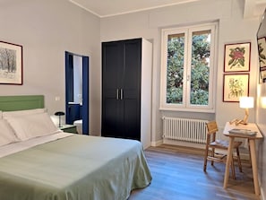 A bedroom with a bed, a nightstand, a desk with a chair, a window with a view of greenery, and a radiator. - St.Peter's Basilica Apartment (Roma)