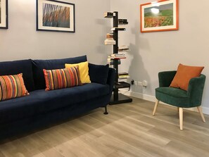 Uma sala de estar com um sofá azul escuro, uma almofada listrada, uma poltrona verde e uma estante contra a parede.