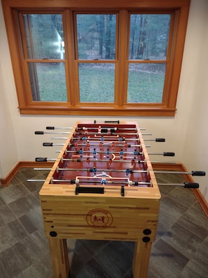 A foosball table with a wooden base and a logo, positioned in front of a window with a view of a grassy area.