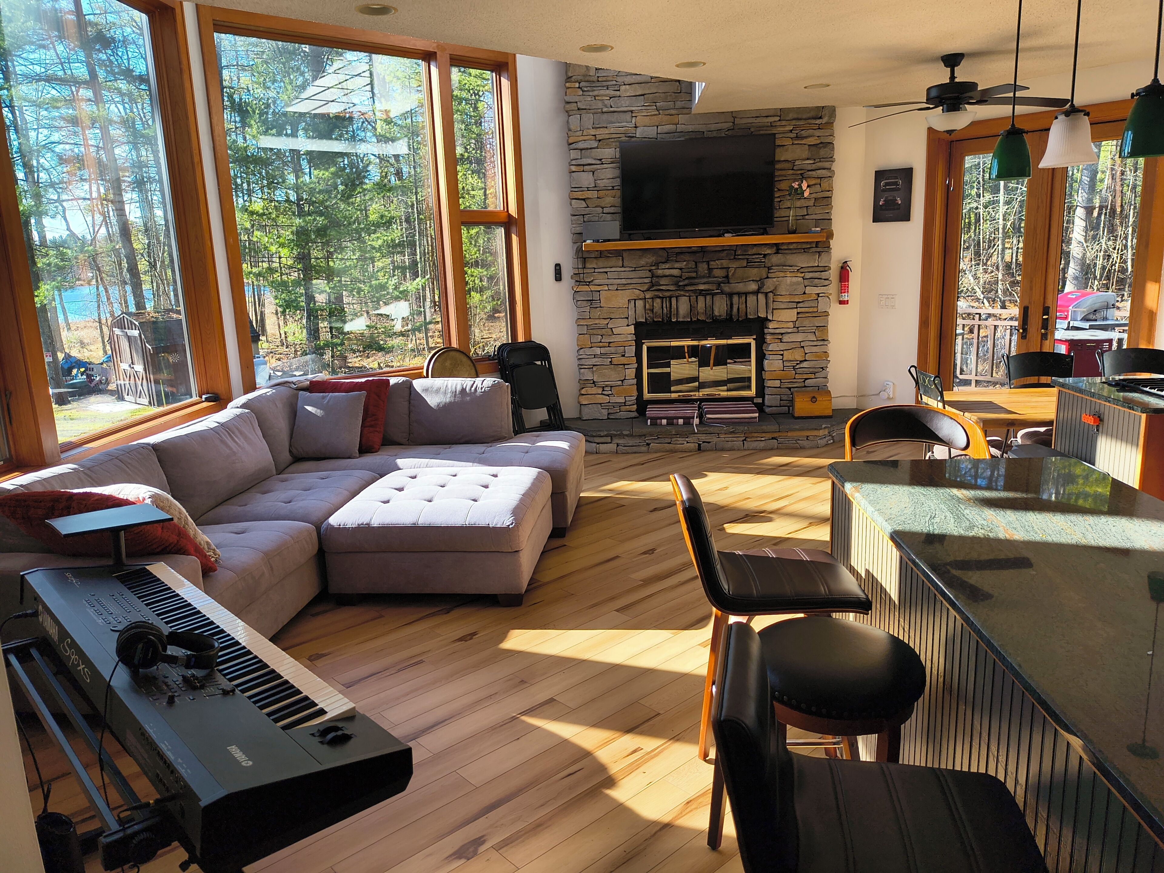 Ein Wohnzimmer mit Ecksofa, einem Klavier, einem Kamin aus Stein und großen Fenstern.