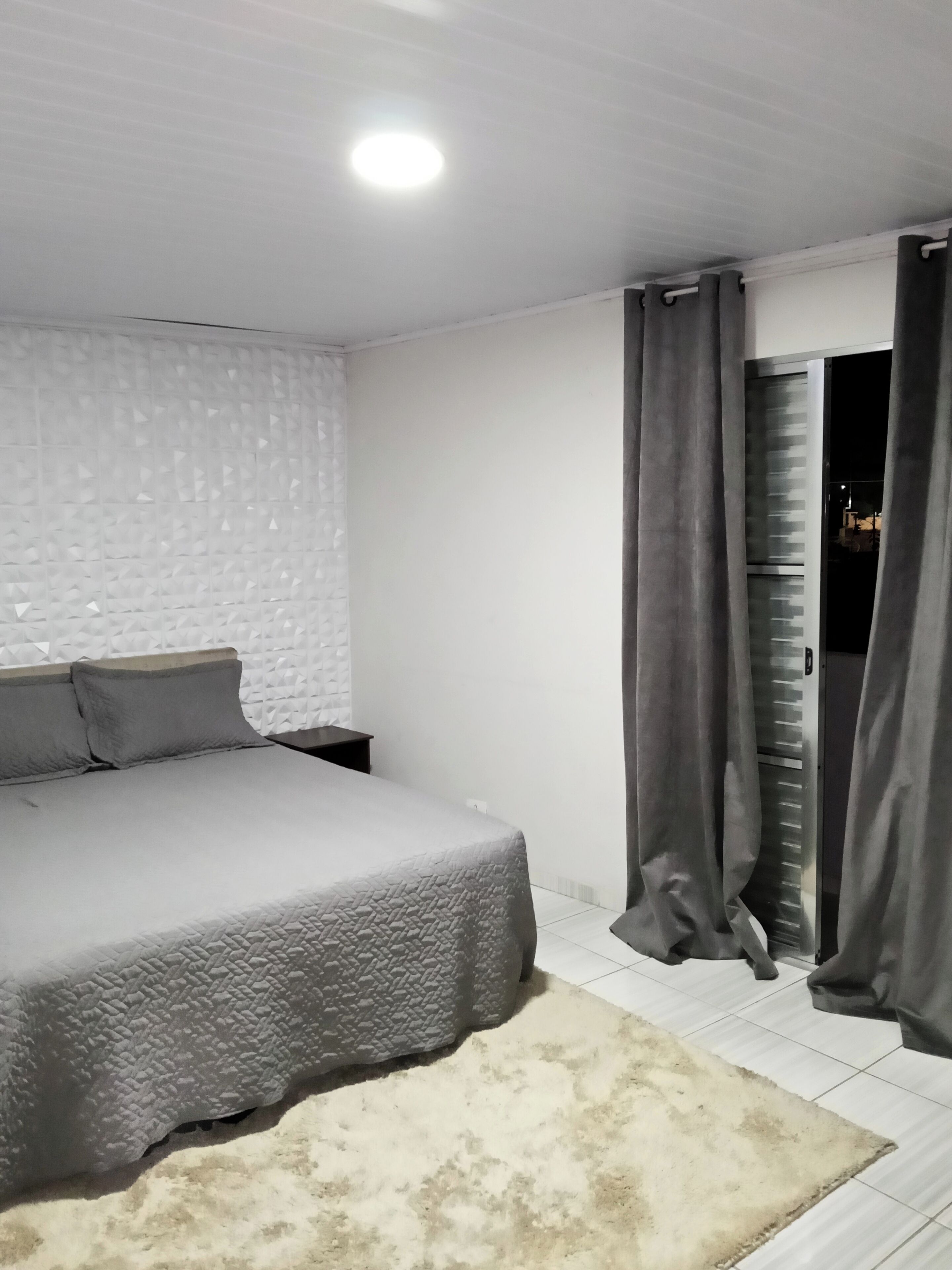 Un dormitorio con una cama, una mesita de noche, una pared blanca con textura y una puerta con cortinas.