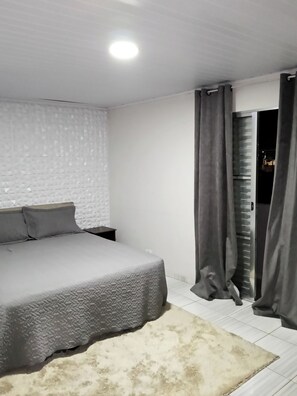 A bedroom with a bed, a nightstand, a white textured wall, and a door with curtains. - Entire house - 19km from Guarulhos Airport (GRU) (Jardim Presidente Dutra)