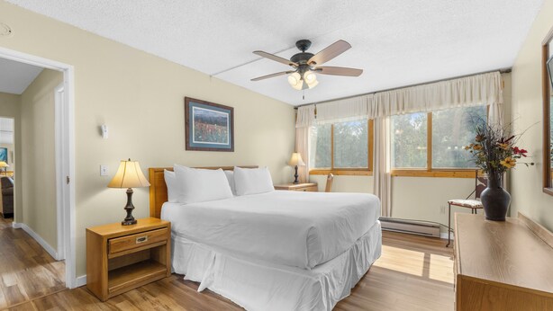 Quarto com cama, mesas de cabeceira, ventilador de teto, janela com cortinas e um quadro na parede.