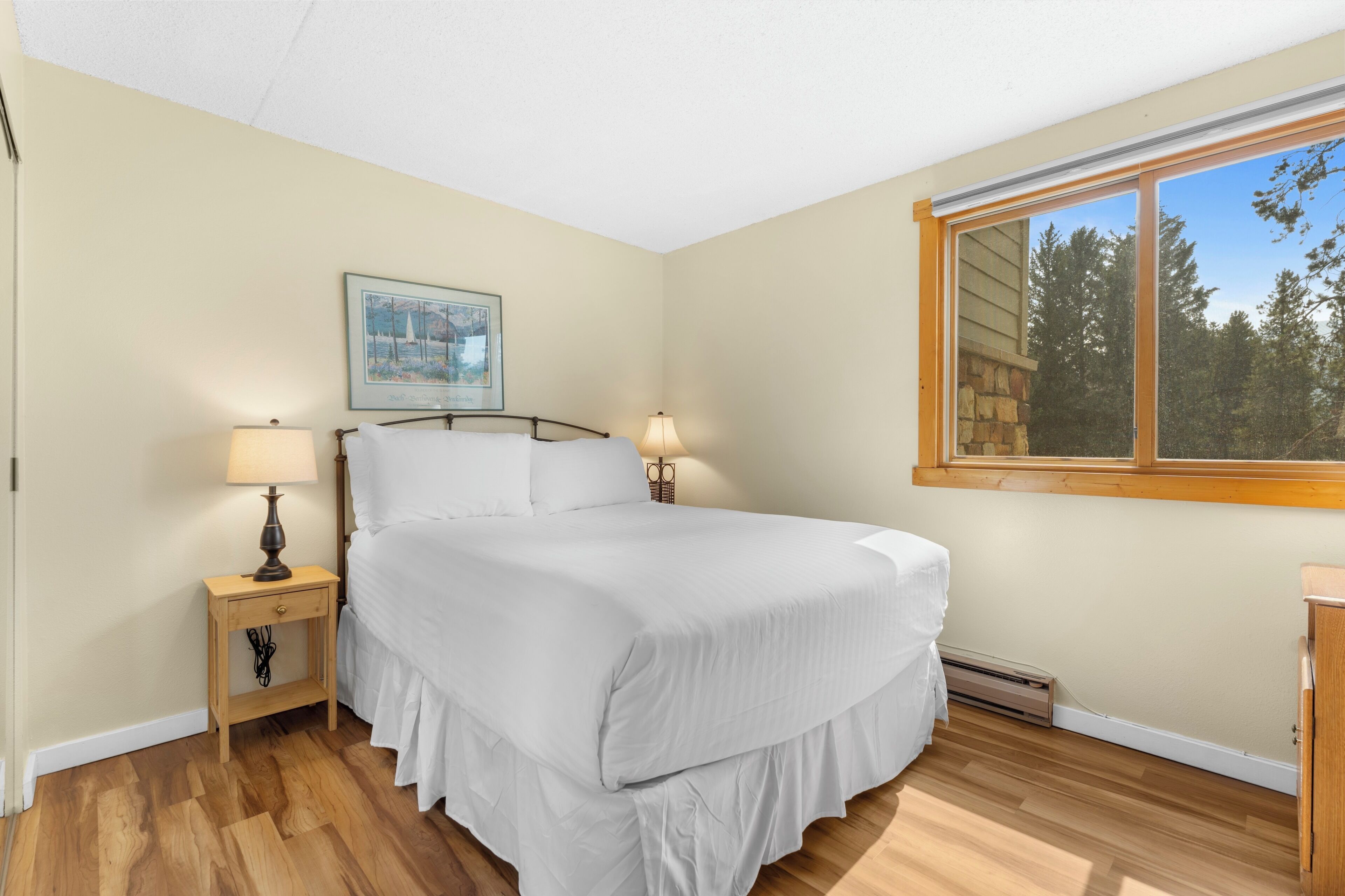 Une chambre à coucher avec un lit, deux tables de chevet, une commode, une fenêtre donnant sur des arbres et un tableau encadré accroché au mur.