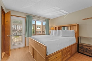 Un dormitorio con cama de madera, ventana con cortinas, una puerta y una cómoda de madera.