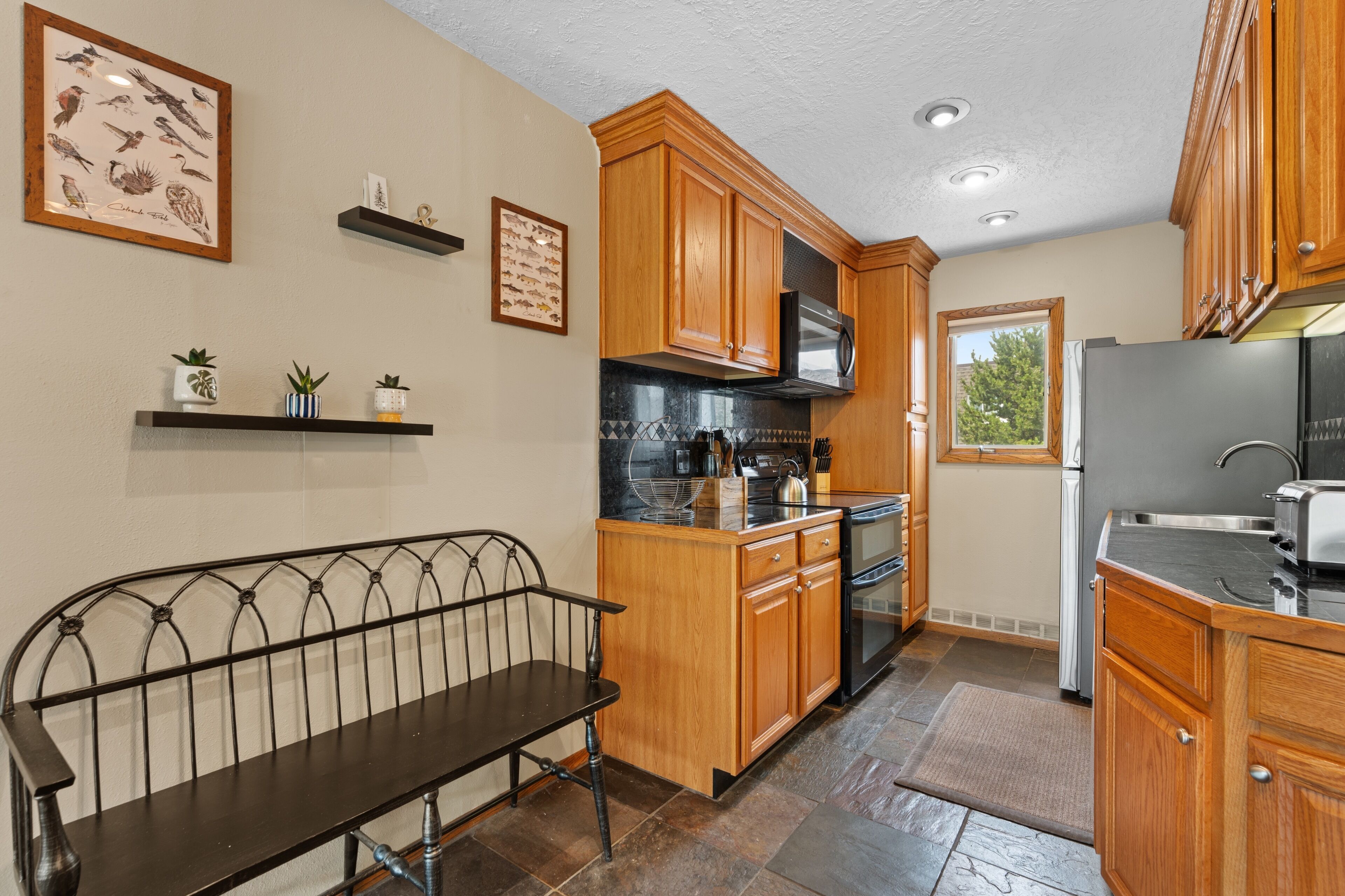 Una cocina con armarios de madera, encimera negra, refrigerador de acero inoxidable y una ventana.