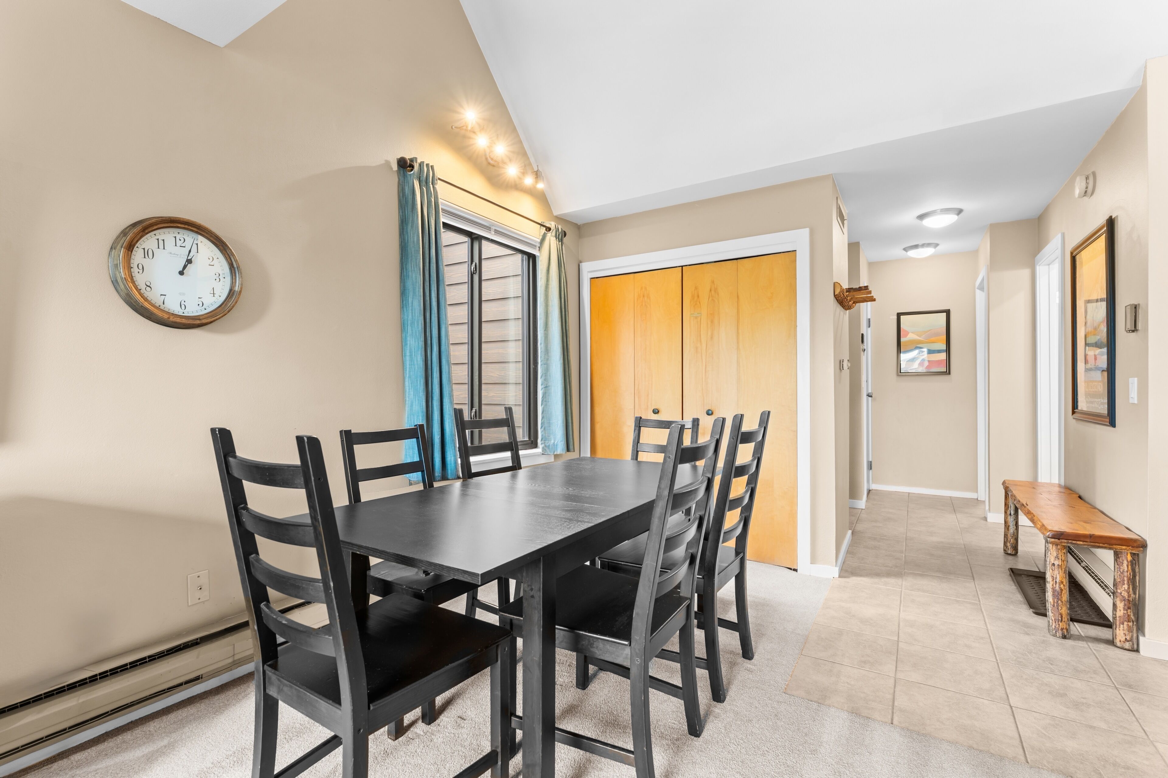Un área de comedor con una mesa y sillas, un reloj en la pared y una puerta que da a otra habitación.