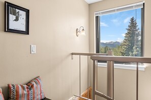 Ein Zimmer mit einem großen Fenster, einem gerahmten Bild an der Wand, einer Lampe und einem gemusterten Kissen.
