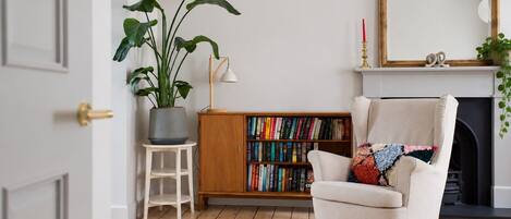 Una habitación con piso de madera, una estantería, un sillón, una planta en maceta, una lámpara y una chimenea.