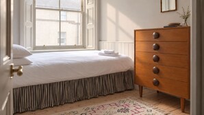 A bedroom with a bed, a dresser, a window with blinds, a mirror, and a patterned rug.