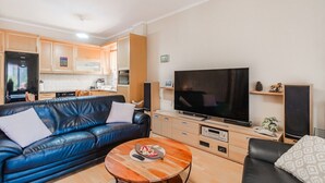 Ein Wohnzimmer mit einem blauen Ledersofa, einem Couchtisch, einem Flachbildfernseher und einer Küche im Hintergrund.