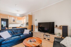 A living room with a blue leather sofa, a coffee table, a flat-screen TV, and a kitchen in the background. - Tilias House by Rentals Pro - Nea Flogita Halkidiki (Nea Flogita)