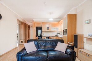 A modern living room with a black leather sofa, wooden cabinets, and a kitchen area in the background. - Tilias House by Rentals Pro - Nea Flogita Halkidiki (Nea Flogita)