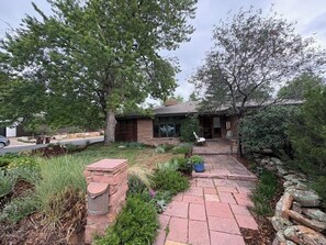 A residential property with a paved driveway leading to a wooden house, surrounded by mature trees and landscaped garden beds. - Sharp Mid-Century Modern Home with Hot Tub -- Old North Boulder (Boulder)