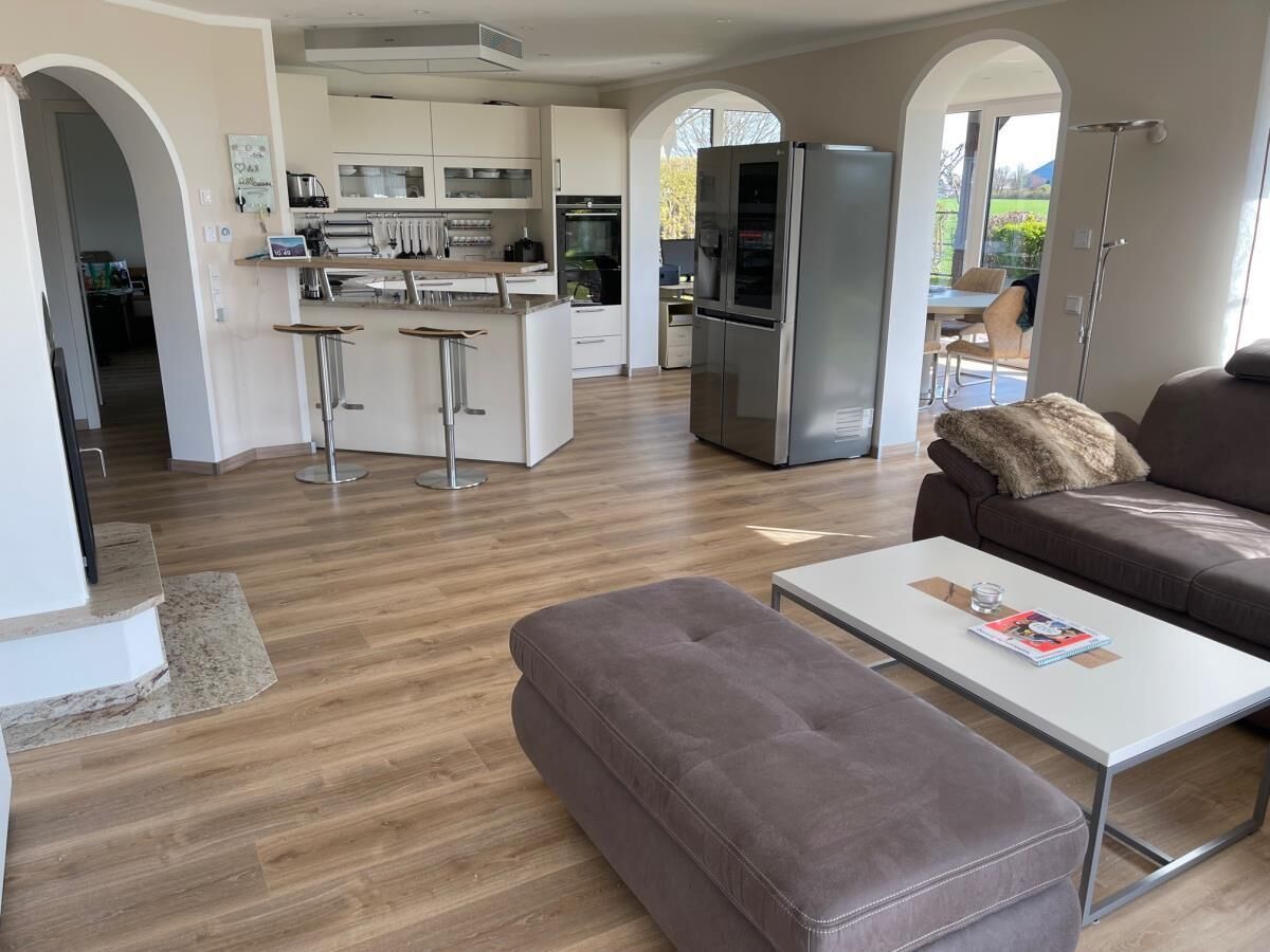 Ein modernes Wohnzimmer mit Sofa, Hocker und Couchtisch. Im Hintergrund ist eine Küche mit Kühlschrank und Barhocker zu sehen.
