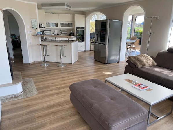 A modern living room with a sofa, ottoman, and coffee table. A kitchen with a refrigerator and bar stools is visible in the background. - 30 Vacation Rental "Anders Fewo 4" (Burg auf Fehmarn)