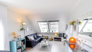 Ein modernes Wohnzimmer mit einer Couch, einem Couchtisch und einem Bücherregal. Zwei Fenster sorgen für natürliches Licht.