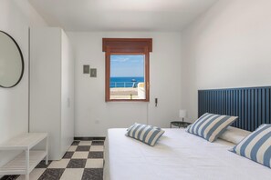 A bedroom with a bed, a bedside table, a nightstand, a window with a view of the sea, and a door.