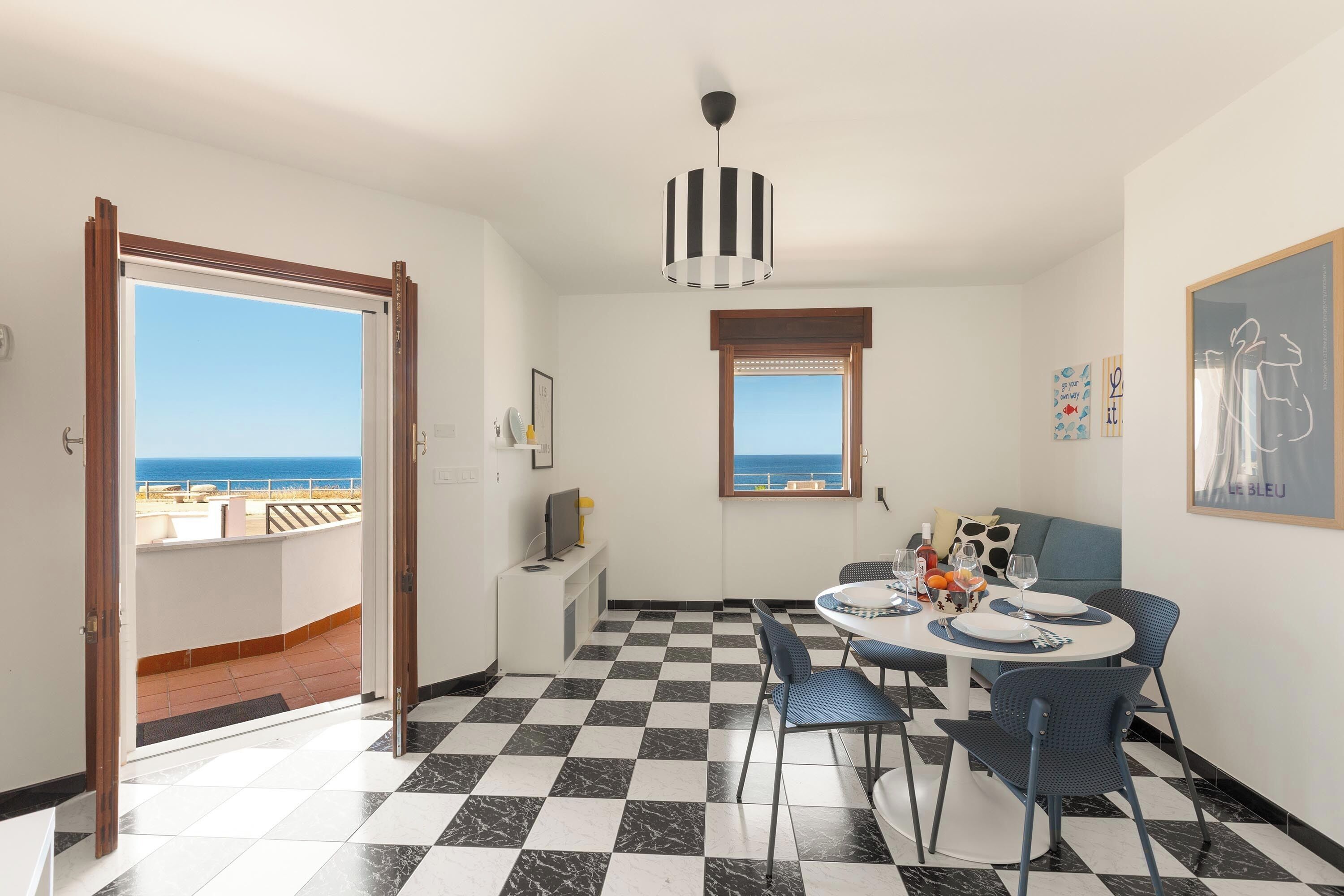 A dining area with a checkered floor, a table set for four, and a view of the sea through a window.