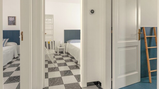 Una habitación de hotel con piso a cuadros, una cama con cabecera a rayas, una mesita de noche con lámpara y una puerta con una escalera de madera apoyada.