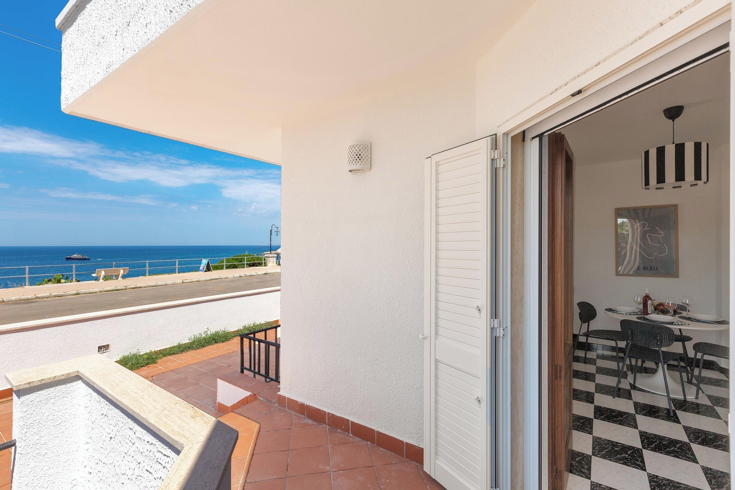 Een balkon met uitzicht op zee, een eethoek met een zwart-wit geruite vloer, en een witte deur.
