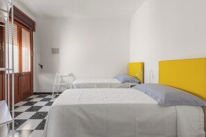 A room with two beds, a white chair, a wooden door, and a checkered floor. - Casa Tramonti - Trilo Superior by BarbarHouse (Santa Maria di Leuca)
