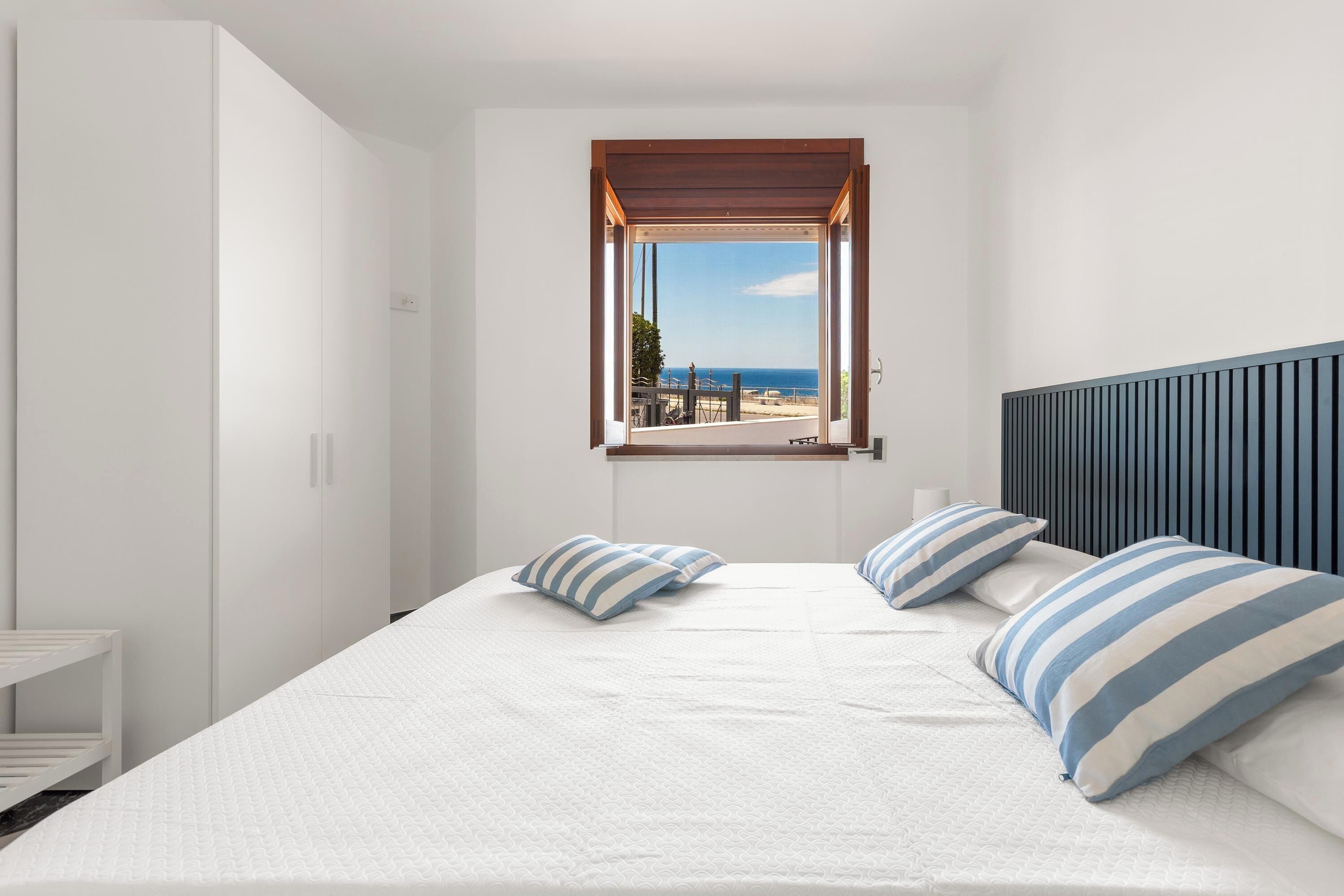 A bedroom with a large bed, a window with a view of the beach, and a white wardrobe.