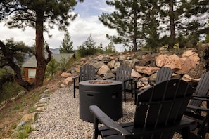 An outdoor seating area with Adirondack chairs and a fire pit, surrounded by rocks and trees. - Log Cabin Retreat - Hot Tub · Sauna · Game Room · Stunning Mountain Views (Pine)
