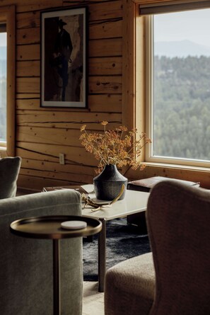 A cozy room with a wooden wall, a framed artwork, a vase with dried flowers, and a view of a forest through a window. - Log Cabin Retreat - Hot Tub · Sauna · Game Room · Stunning Mountain Views (Pine)