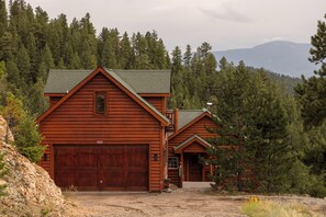 A two-story log cabin with a large garage, surrounded by dense forest and mountains in the background. - Log Cabin Retreat - Hot Tub · Sauna · Game Room · Stunning Mountain Views (Pine)