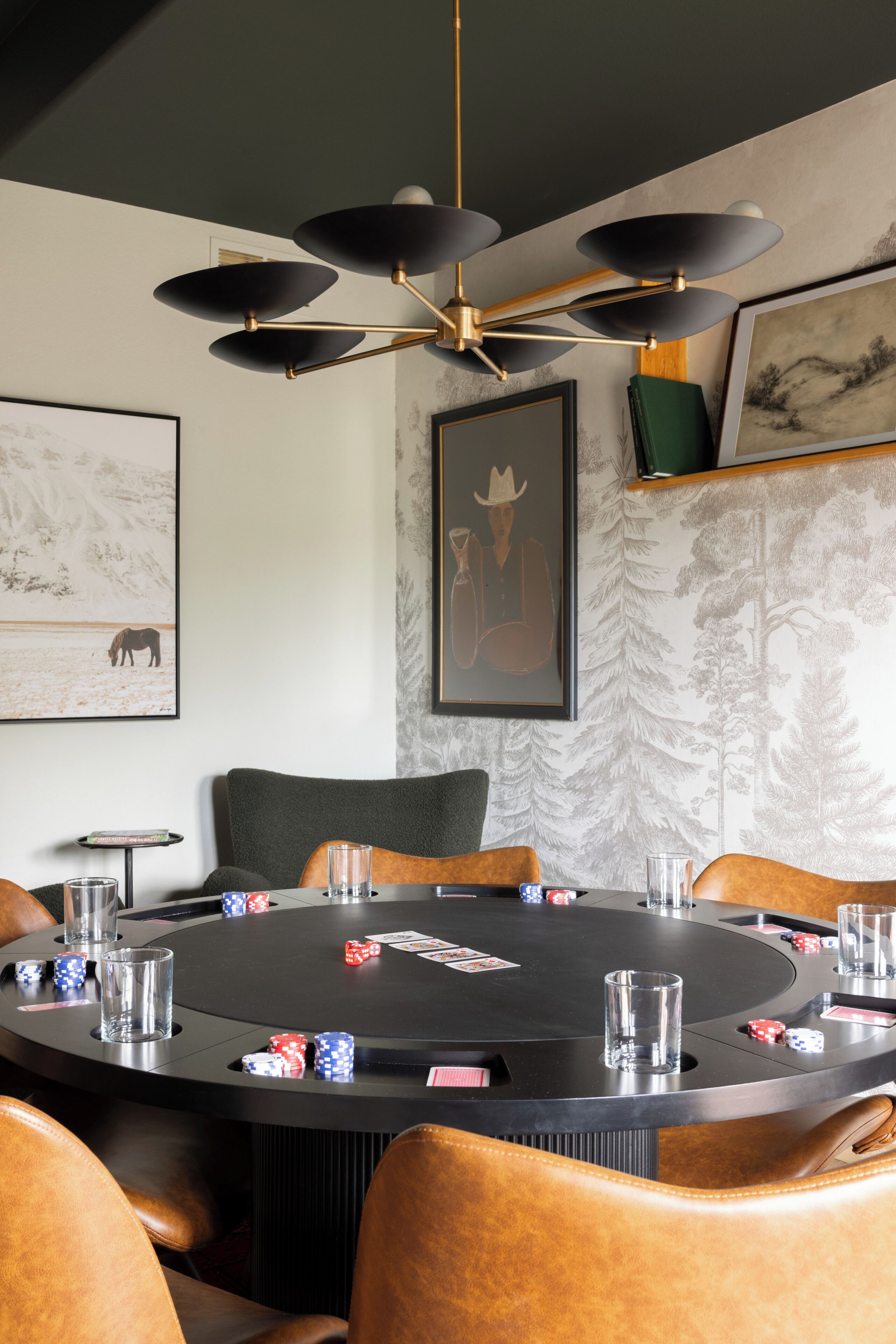 Una mesa redonda con fichas de póker y cartas, rodeada de sillones de cuero, en una habitación con ventilador de techo y cuadros en las paredes.