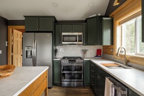 A modern kitchen with green cabinets, stainless steel appliances, and a large window providing natural light. - Log Cabin Retreat - Hot Tub · Sauna · Game Room · Stunning Mountain Views (Pine)