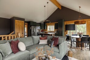 A modern living room with a sectional sofa, a dining area with a table and chairs, and a kitchen with dark cabinets and a large window. - Log Cabin Retreat - Hot Tub · Sauna · Game Room · Stunning Mountain Views (Pine)