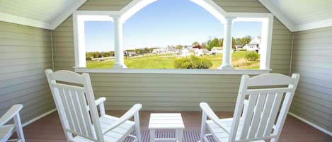 A covered porch with two white Adirondack chairs and a small table, overlooking a residential area.
