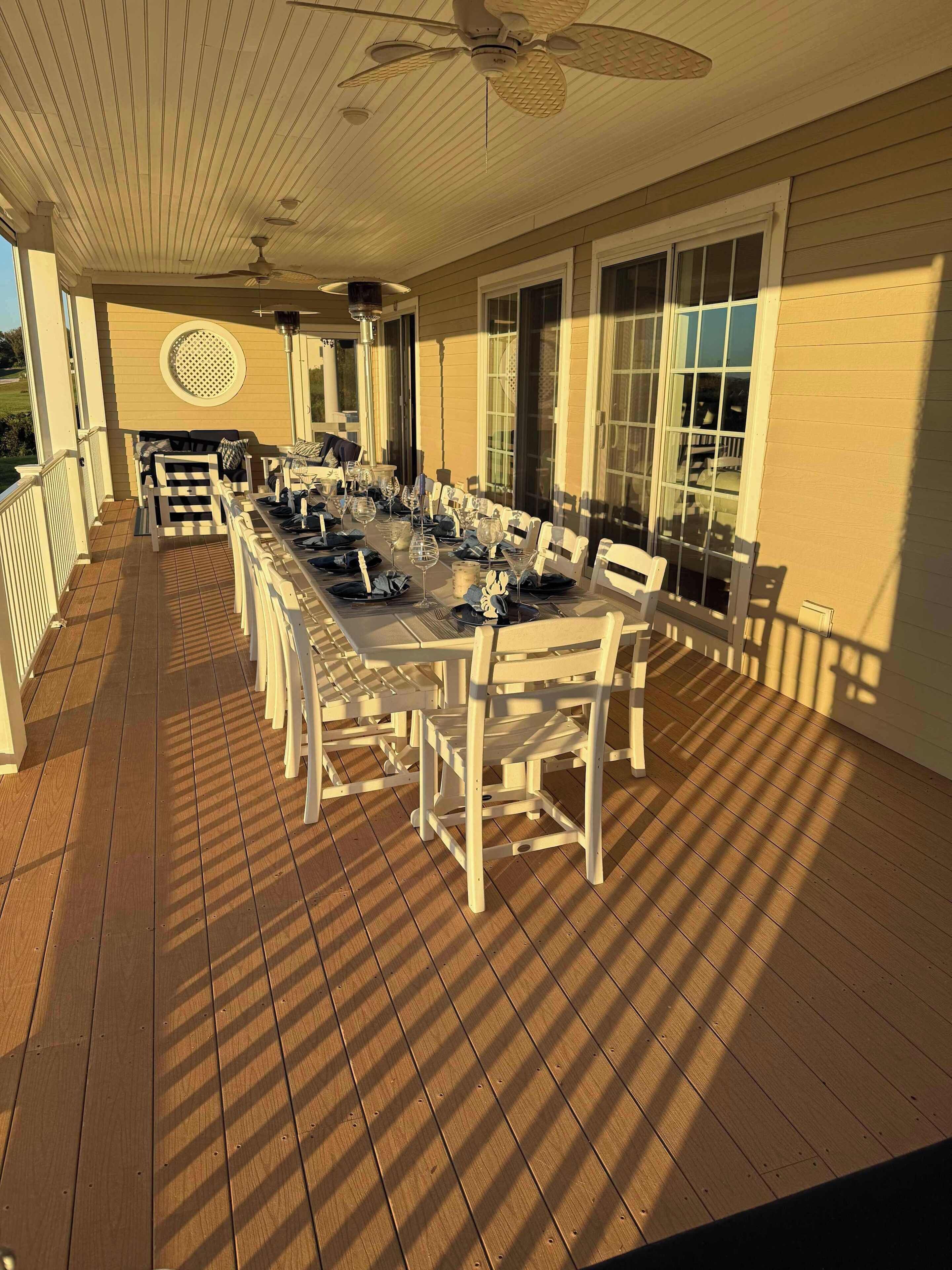A covered outdoor dining area with a long table set for multiple guests, surrounded by white chairs, and a ceiling fan overhead.