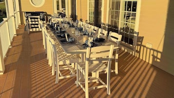 A covered outdoor dining area with a long table set for multiple guests, surrounded by white chairs, and a ceiling fan overhead.