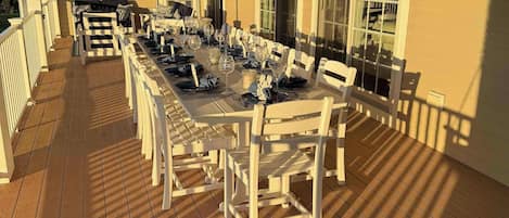 A covered outdoor dining area with a long table set for multiple guests, surrounded by white chairs, and a ceiling fan overhead.