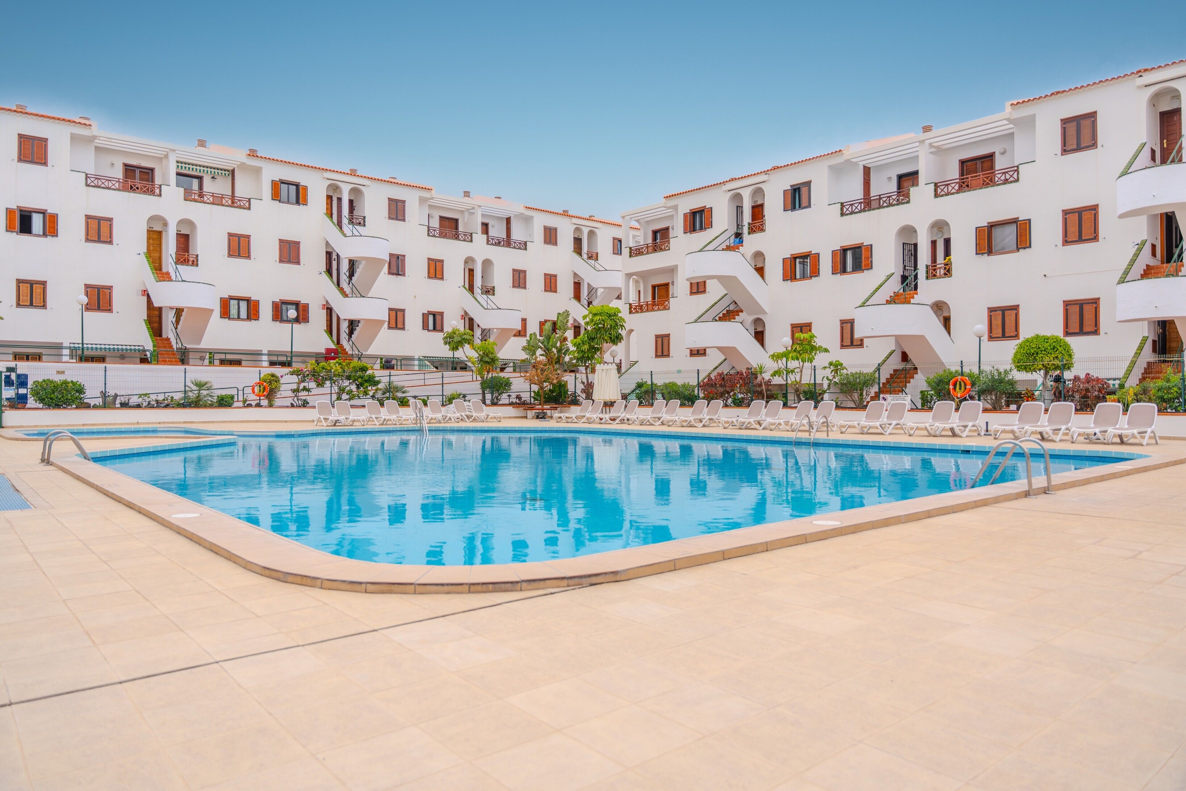 Une piscine entourée de bâtiments blancs dotés de balcons, sous un ciel dégagé.