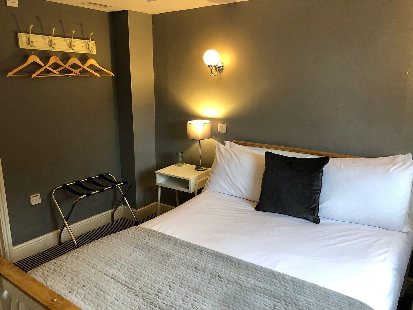 Habitación de hotel con cama, mesita de noche con lámpara, perchero y una silla.