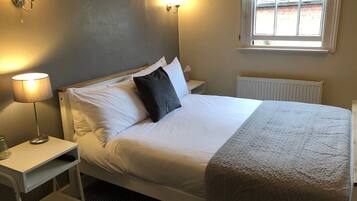A neatly made bed with white linens and a grey quilt, a bedside lamp, a nightstand with a glass, and a window with blinds.