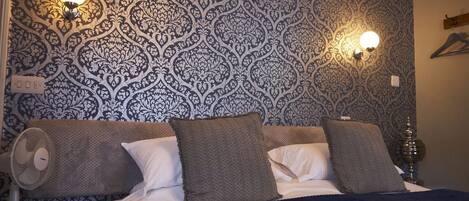 A bedroom with a patterned wallpaper, two wall-mounted lamps, a bed with pillows, a bedside table with a lamp, and a fan.