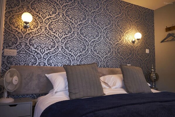 A bedroom with a patterned wallpaper, two wall-mounted lamps, a bed with pillows, a bedside table with a lamp, and a fan. - Coachmakers Arms (Wallingford)