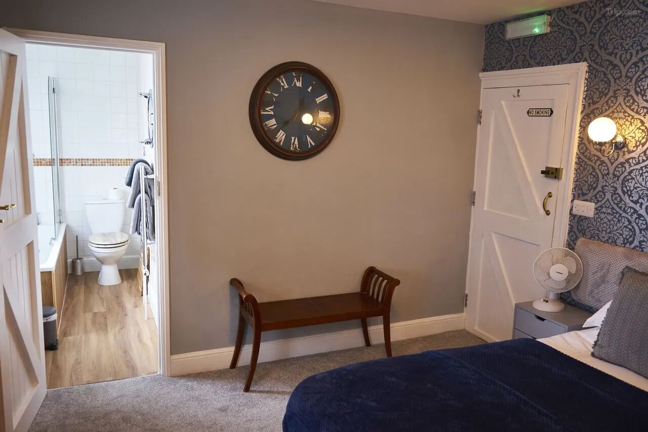 A bedroom with a bed, a bedside table with a fan, a wall clock, a bench, and a door leading to a bathroom.