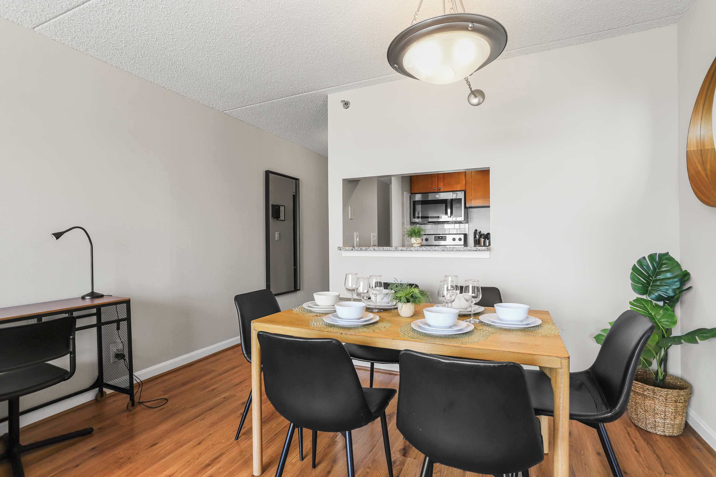 A dining area with a wooden table set for six, black chairs, a hanging light fixture, and a kitchen visible through an open door.